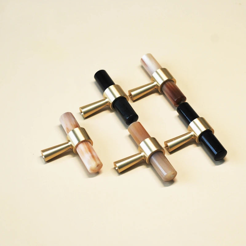 vijf T-model meubelknopjes mat messing met een staafje van hoorn zwart en gemêleerd wit  liggend op een beige ondergrond 