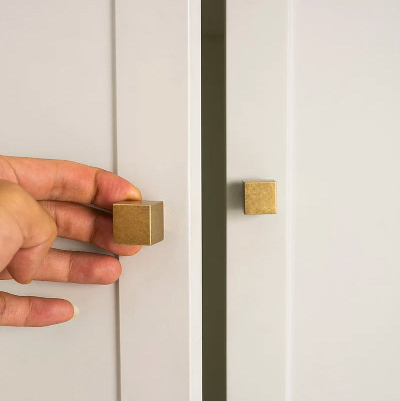 twee vierkante antiek messing meubelknopjes gemonteerd op een witte kast een hand pakt 1 knopje